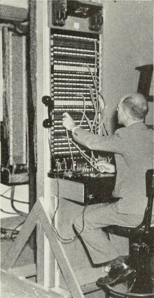 White man operates a complicated switchboard with a mess of protruding wires