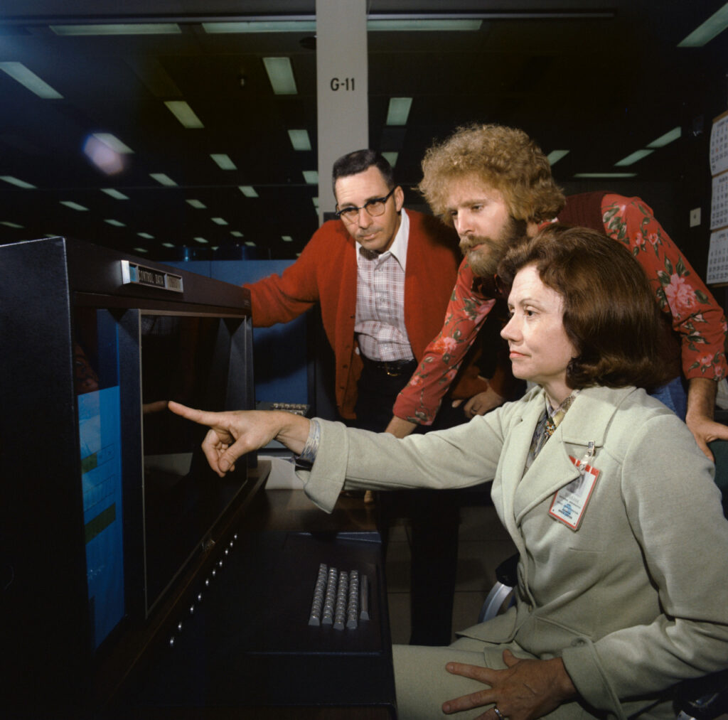 Three white people look at a computer screen. A seated woman points on the monitor